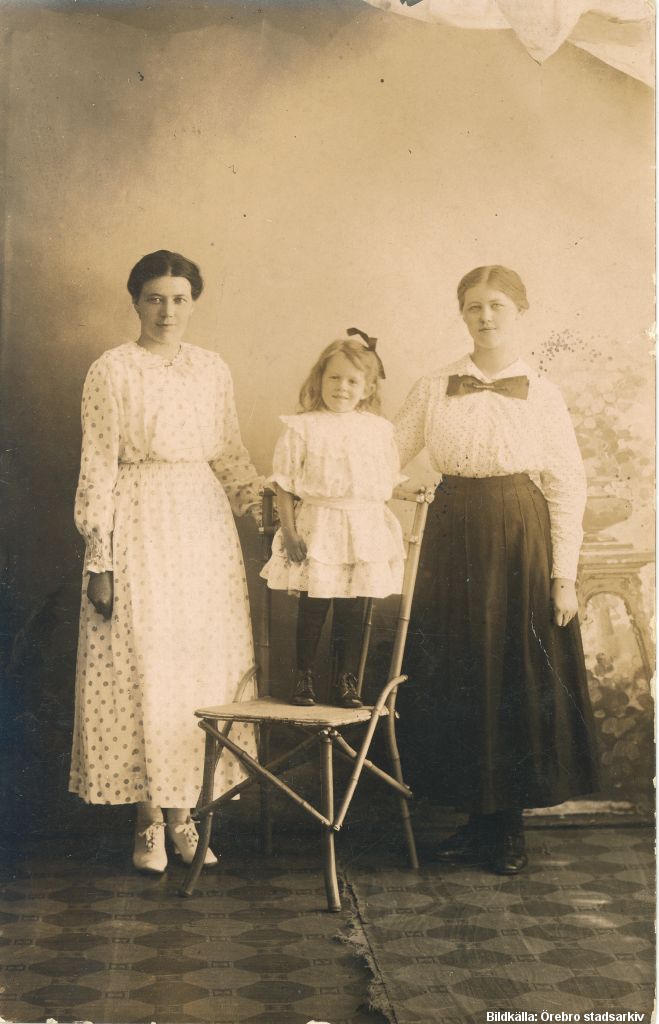 En flicka står på en stol mellan två kvinnor i en fotoateljé.