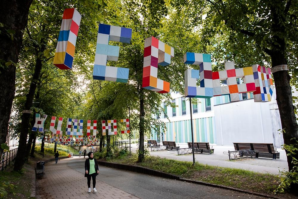 Above a bicycle lane in the trees hang large letters from the Latin, Hebrew and Cyrillic alphabets. The letters are three-dimensional, white and with sewn-on rectangular textile pieces in blue, red and orange tones.